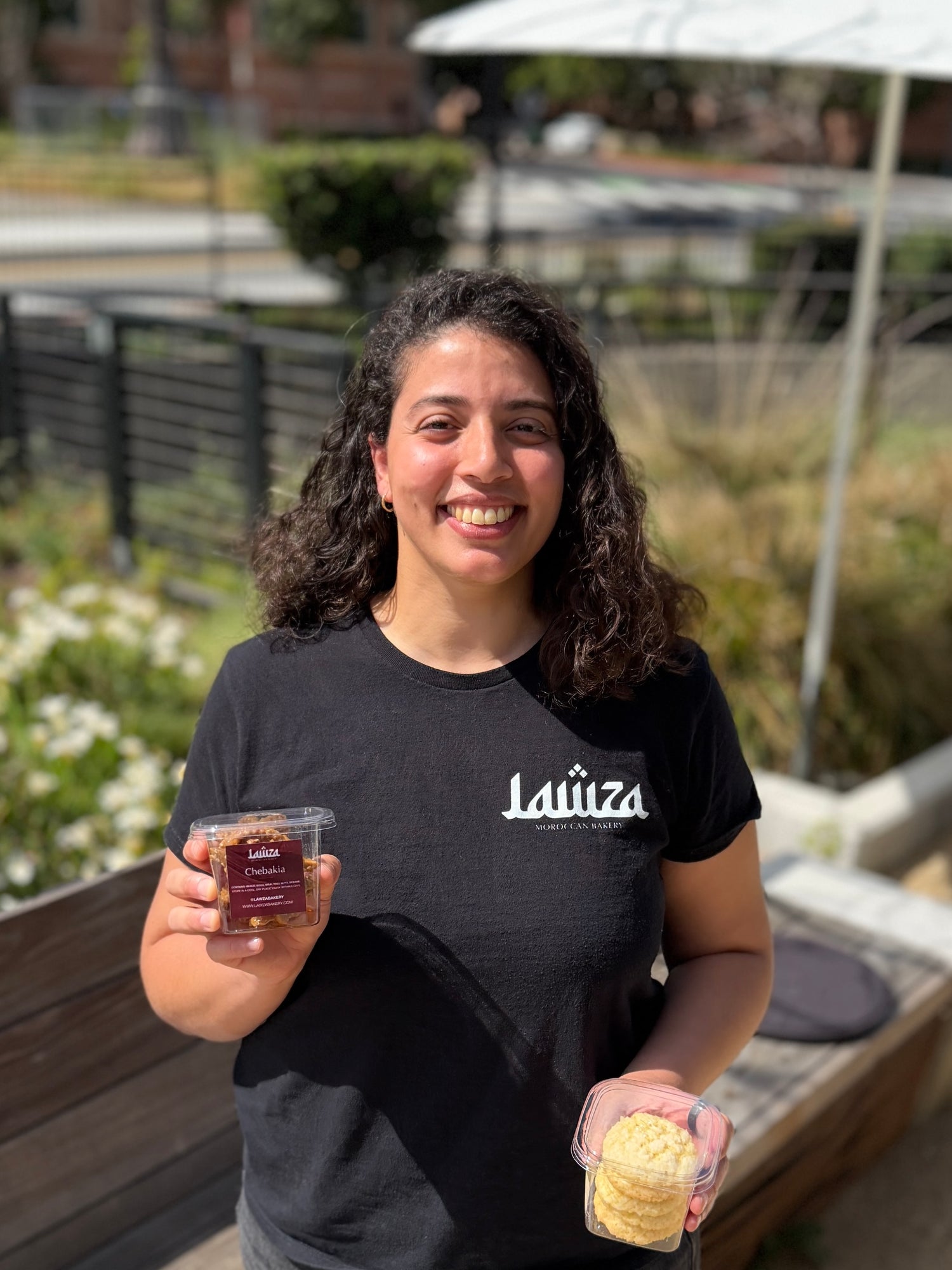 Founder of Lawza Bakery, holding cups of Moroccan sweets and wearing a Lawza branded t-shirt.