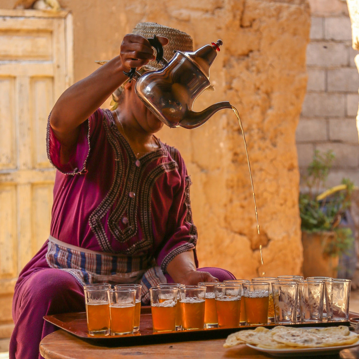 Person serving tea from a traditional teapot in an outdoor setting with stone walls and wooden furniture.