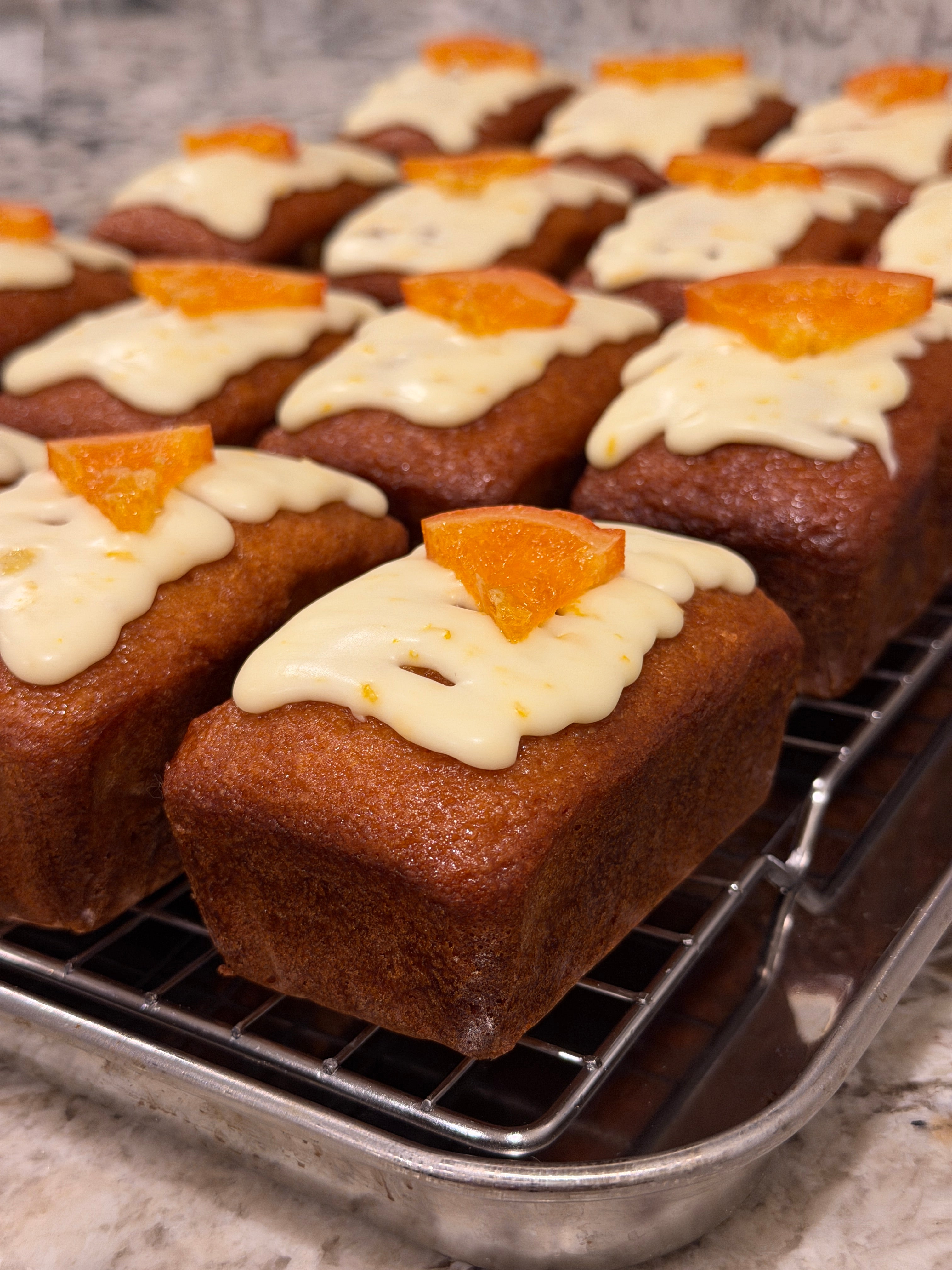 Row of Moroccan orange mini cakes with white frosting and orange pieces on a metal cooling rack
